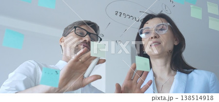 Business colleagues collaborating on strategy, brainstorming ideas for success, writing on a transparent glass board with sticky notes in a modern office 138514918