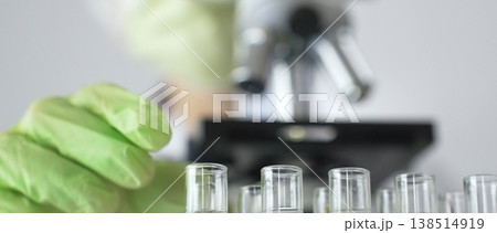Scientist wearing green gloves and a face mask selecting a test tube with transparent liquid from a rack near a microscope in a lab. Medicine, healthcare and science concept Scientist wearing green gloves and a face mask selecting a test tube with transparent liquid from a rack near a microscope in a lab. Medicine, healthcare and science concept 138514919