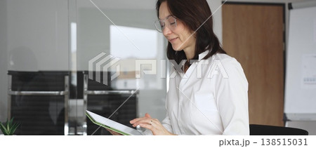 Businesswoman in white blouse managing tasks with a digital tablet, sitting at a modern glass desk in a bright office environment, engaging in remote work, online learning, and financial analysis Businesswoman in white blouse managing tasks with a digital tablet, sitting at a modern glass desk in a bright office environment, engaging in remote work, online learning, and financial analysis 138515031