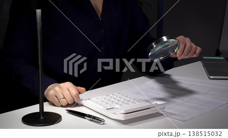 Person checking financial documents and numbers with a magnifying glass, analyzing business data for budgeting and taxes at night. Audit and taxes in business Person checking financial documents and numbers with a magnifying glass, analyzing business data for budgeting and taxes at night. Audit and taxes in business 138515032