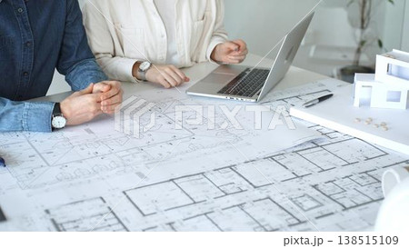 Architects engaging in a professional meeting, reviewing detailed blueprints on a table, utilizing a laptop, and collaborating on building design and construction planning 138515109