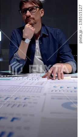 Professional man in glasses thinking deeply while analyzing financial charts and data on an office desk, working late night in a dark setting, feeling focused and determined 138515127