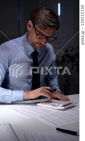 Accountant working late night in office, calculating finance data and economy statistics in a blue shirt with a tie. Audit and taxes in business Accountant working late night in office, calculating finance data and economy statistics in a blue shirt with a tie. Audit and taxes in business 138515130