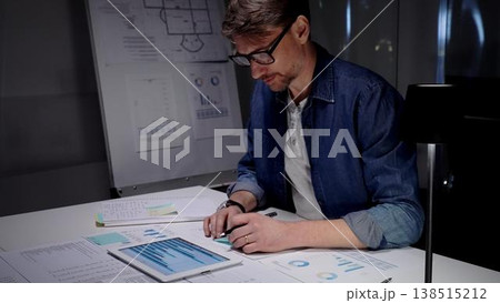 Professional man in glasses working late at night, analyzing financial data on a digital tablet, managing business reports, and extending productivity in a dark office environment 138515212