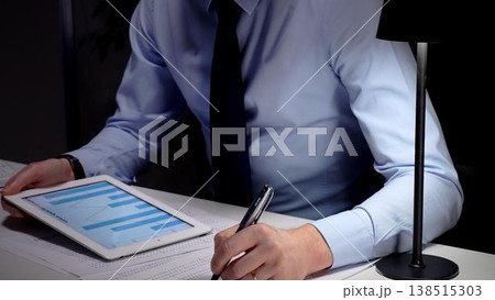Businessman working late analyzing financial data at office, holding tablet with charts, writing notes on documents. Audit and taxes in business Businessman working late analyzing financial data at office, holding tablet with charts, writing notes on documents. Audit and taxes in business 138515303