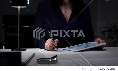 Woman working late night in office, multitasking with a tablet and writing on documents, managing heavy paperwork load. Audit and taxes in business 138515360