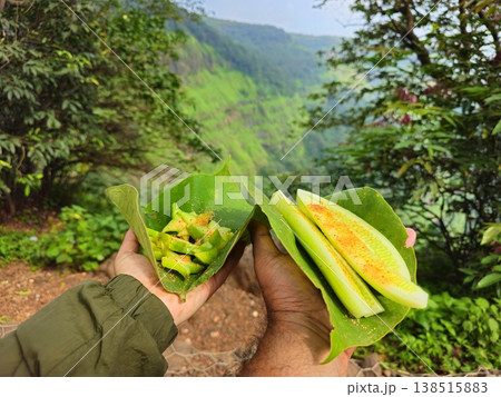 Hand holding sliced cucumber and fruit snacks on leaves against a lush green mountain backdrop, combining travel, food, and nature in Matheran. 138515883