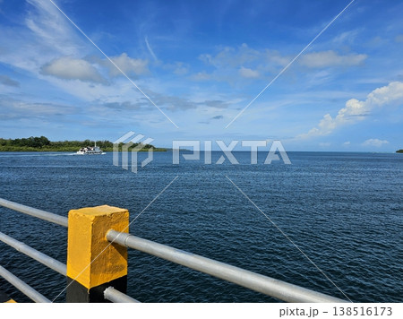 Scenic coastal view from a pier featuring yellow and black concrete bollards, looking out over calm ocean waters towards a distant green island. Scenic coastal view from a pier featuring yellow and black concrete bollards, looking out over calm ocean waters towards a distant green island. 138516173