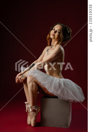 Modern tattooed ballerina in a white tutu and pointe shoes posing on a burgundy background. 138519338