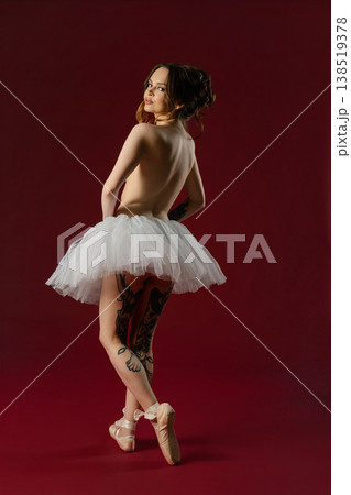 Elegant tattooed ballerina in a white tutu and pointe shoes looking back over her shoulder against a red background. Elegant tattooed ballerina in a white tutu and pointe shoes looking back over her shoulder against a red background. 138519378