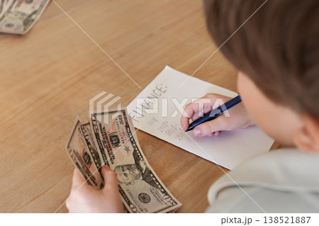 Child counting US dollar cash and writing finance notes on paper on beige wooden table. Early financial literacy, budgeting skills and money awareness concept with copy space indoors Child counting US dollar cash and writing finance notes on paper on beige wooden table. Early financial literacy, budgeting skills and money awareness concept with copy space indoors 138521887