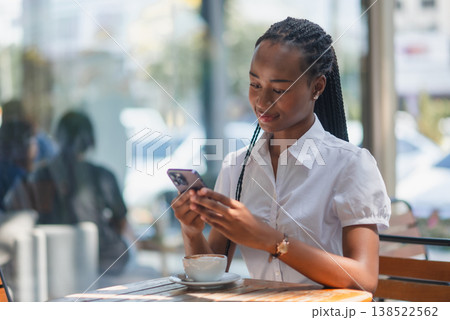Young Black woman using smartphone at cafe table with coffee cup, smiling during a casual work break. Modern mobile lifestyle, online communication, remote work and everyday technology concept. Young Black woman using smartphone at cafe table with coffee cup, smiling during a casual work break. Modern mobile lifestyle, online communication, remote work and everyday technology concept. 138522562