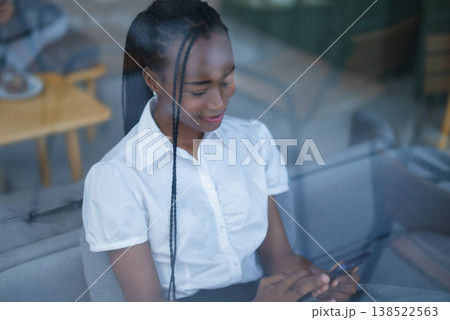 Young Black woman using smartphone behind window glass, seated on a sofa with soft reflections. Candid digital lifestyle, indoor communication, mobile browsing and modern connection. Young Black woman using smartphone behind window glass, seated on a sofa with soft reflections. Candid digital lifestyle, indoor communication, mobile browsing and modern connection. 138522563