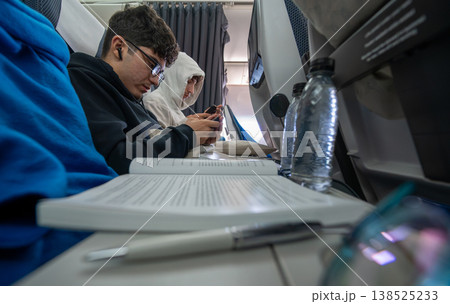 Airplane passengers sitting on comfortable seats, a young man wearing glasses looking at his smartphone, another person using a phone, and a book with a pen on the tray table during a flight 138525233