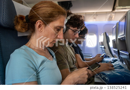 Passengers seated in an airplane cabin focus on their mobile devices during a flight, showing modern technology use and digital connectivity while traveling together Passengers seated in an airplane cabin focus on their mobile devices during a flight, showing modern technology use and digital connectivity while traveling together 138525241