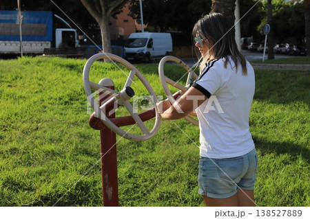 Young woman exercises on outdoor fitness equipment in a park, surrounded by green grass and trees, with vehicles visible in the background 138527889