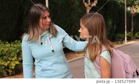 Mom and young daughter stand together outdoors near school building, girl with backpack talking animatedly 138527899