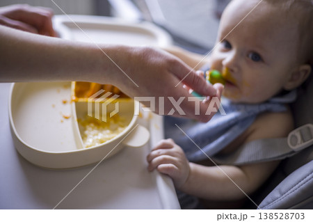 White Infant Hesitant To Accept Spoon, Parent Hand Reaching Across Tray, Messy Bib And Bright Orange Puree, Quiet Tension And Patient Encouragement In Home Feeding Scene 138528703