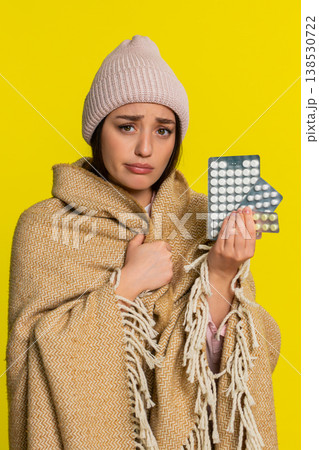 Woman wrapped in blanket wearing hat holding pills thoughtfully looking at them cold flu treatment 138530722