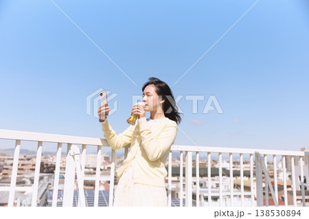 屋外でスマートフォンを持ちながら飲み物を楽しむ女性 青空の下で自撮りするライフスタイル 138530894