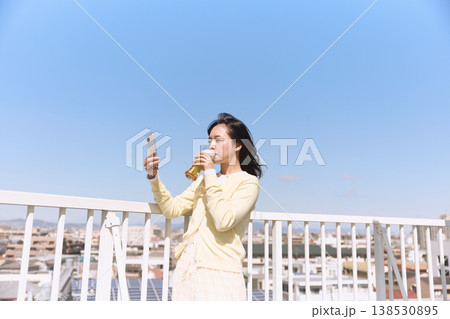 屋外でスマートフォンを持ちながら飲み物を楽しむ女性 青空の下で自撮りするライフスタイル 屋外でスマートフォンを持ちながら飲み物を楽しむ女性 青空の下で自撮りするライフスタイル 138530895