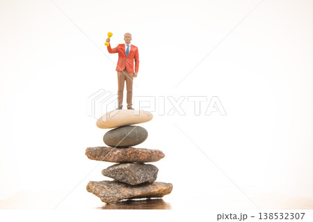 Miniature figurine stands on stones holding a trophy in a simple setting with a white background Miniature figurine stands on stones holding a trophy in a simple setting with a white background 138532307
