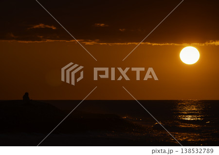 Silhouette of person sitting by sea at intense red sunset with bright sun and reflection on water, dramatic sky and waves, emotional seascape with copy space, solitude and contemplation concept Silhouette of person sitting by sea at intense red sunset with bright sun and reflection on water, dramatic sky and waves, emotional seascape with copy space, solitude and contemplation concept 138532789