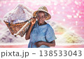 A years old woman stands in a pink salt lake in Senegal. She carries a basket filled with salt on her shoulder. The sun shines brightly on the scene. Salt harvesting is taking place in the background. 138533043
