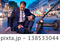 A middle aged businessman in a suit sits on the street, looking shocked and holding his knee. An electric scooter is nearby. The scene shows a busy urban setting during evening rush hour. 138533044