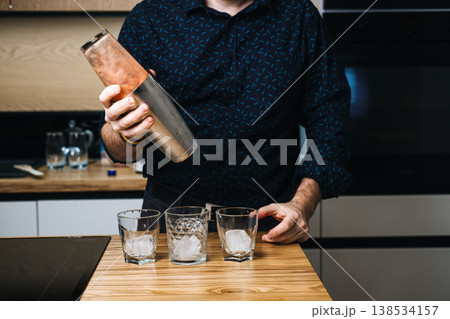 Man shakes metal cocktail shaker over three glasses filled with ice on wooden counter. Professional bartender, cocktail mixology, craft drink preparation. Man shakes metal cocktail shaker over three glasses filled with ice on wooden counter. Professional bartender, cocktail mixology, craft drink preparation. 138534157