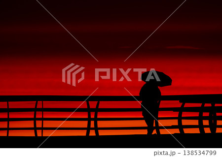 Silhouette of person with umbrella on pier at red sunset over sea, minimal coastal scene with dramatic sky and ocean horizon, evening mood with vivid orange and crimson tones Silhouette of person with umbrella on pier at red sunset over sea, minimal coastal scene with dramatic sky and ocean horizon, evening mood with vivid orange and crimson tones 138534799