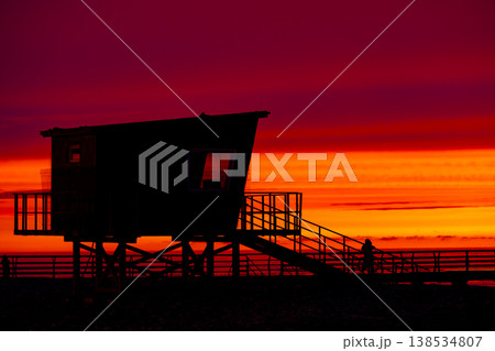 Silhouette of lifeguard tower on beach at dramatic red sunset, coastal structure with stairs and pier, ocean horizon in background, minimalistic evening scene with vivid sky and travel mood 138534807