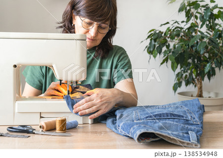 An adult woman repairs and alters denim jeans on a sewing machine at home, focused on an upcycling diy craft project. Reusing things. Environmental awareness. Eco-friendly lifestyle An adult woman repairs and alters denim jeans on a sewing machine at home, focused on an upcycling diy craft project. Reusing things. Environmental awareness. Eco-friendly lifestyle 138534948