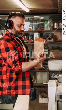 Joinery process. Stacking wood. Bearded carpenter with earmuffs checking freshly cut plank in garage workshop with tools set on wall. Joinery process. Stacking wood. Bearded carpenter with earmuffs checking freshly cut plank in garage workshop with tools set on wall. 138534962