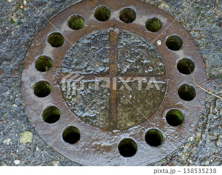 Close-up of a rusty round cast iron manhole cover with drainage holes on wet asphalt. 138535238