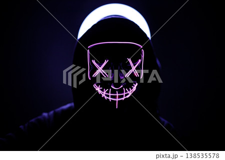 Person in Dark Hood Wearing Glowing Neon Purple Mask with X-Shaped Eyes, Atmospheric Portrait with Backlit Halo Effect, Cyberpunk and Mystery Concept Photography in Total Darkness. 138535578