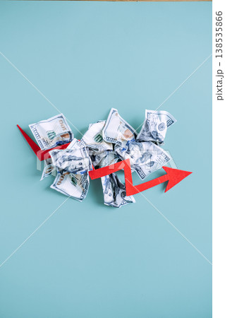 Red arrow points downward over pile of crumpled dollar bills on blue background. Economic recession, deflationary pressure, financial market crash. Red arrow points downward over pile of crumpled dollar bills on blue background. Economic recession, deflationary pressure, financial market crash. 138535866