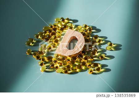 Small wooden letter D stands among yellow translucent supplement capsules on blue surface. Vitamin D deficiency, immune system support, preventative medicine. 138535869