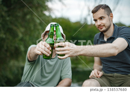 Cropped photo of a men clinking with two bottles of beer. Father and adult son near the lake fishing at summer. Men wearing khaki clothes. Cropped photo of a men clinking with two bottles of beer. Father and adult son near the lake fishing at summer. Men wearing khaki clothes. 138537680