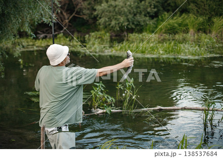 Man with fishing rod near the lake fishing at summer. Fisherman catch a fish. Man wearing khaki clothes and a cap. Man with fishing rod near the lake fishing at summer. Fisherman catch a fish. Man wearing khaki clothes and a cap. 138537684