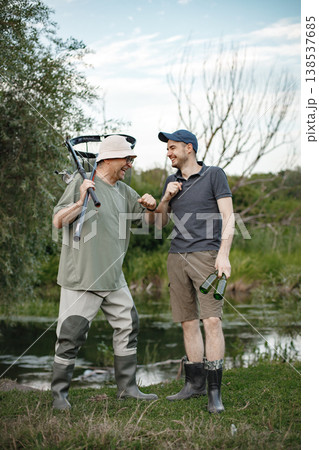Father and adult son with fishing rods near the lake fishing at summer. Fishermen talking together. Men wearing khaki clothes and a caps. 138537685