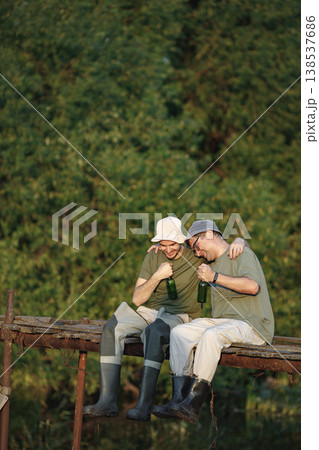Father and adult son with fishing rods near the lake fishing at summer. Fishermen sitting on a footbridge and drinking a beer. Men wearing khaki clothes and a caps. Father and adult son with fishing rods near the lake fishing at summer. Fishermen sitting on a footbridge and drinking a beer. Men wearing khaki clothes and a caps. 138537686