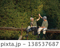 Father and adult son with fishing rods near the lake fishing at summer. Fishermen sitting on a footbridge. Men wearing khaki clothes and a caps. 138537687