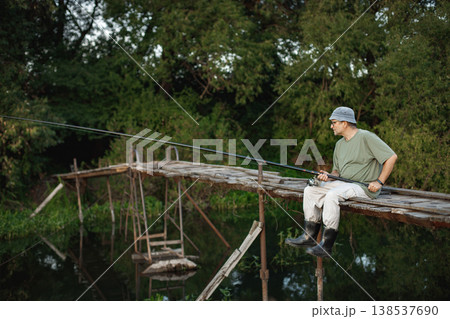 Man with fishing rod near the lake fishing at summer. Fisherman sitting on a footbridge. Man wearing khaki clothes and a cap. Man with fishing rod near the lake fishing at summer. Fisherman sitting on a footbridge. Man wearing khaki clothes and a cap. 138537690