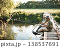 Two men with fishing rods near the lake fishing at summer. Fishermen sitting on a footbridge. Men wearing khaki clothes and a caps. 138537691