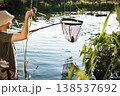 Man with fishing rod and net near the lake fishing at summer. Fisherman enjoying his hobby. Man wearing khaki clothes and a cap. 138537692