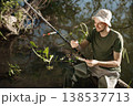Man with fishing rod near the lake fishing at summer. Fisherman enjoying his hobby. Man wearing khaki clothes and a cap. 138537713