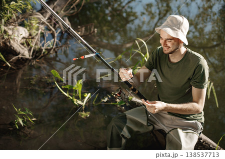 Man with fishing rod near the lake fishing at summer. Fisherman enjoying his hobby. Man wearing khaki clothes and a cap. 138537713