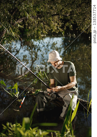 Man with fishing rod near the lake fishing at summer. Fisherman enjoying his hobby. Man wearing khaki clothes and a cap. Man with fishing rod near the lake fishing at summer. Fisherman enjoying his hobby. Man wearing khaki clothes and a cap. 138537714