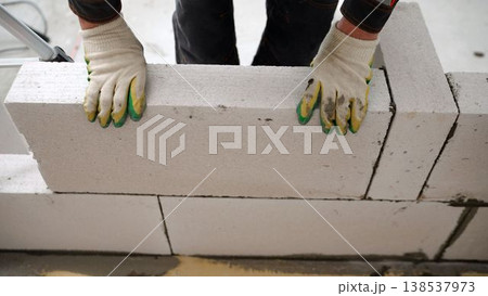 Construction worker in gloves placing a large aerated concrete block while building a wall at a construction site. Construction worker in gloves placing a large aerated concrete block while building a wall at a construction site. 138537973